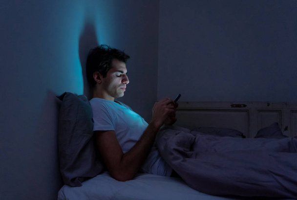 Lifestyle portrait of cell phone addict man awake late at night in bed using smart phone checking likes and followers, chatting, flirting, dating on social media. Internet addiction and mobile abuse. - 写真・画像