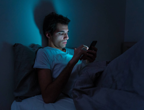 Lifestyle portrait of cell phone addict man awake late at night in bed using smart phone checking likes and followers, chatting, flirting, dating on social media. Internet addiction and mobile abuse. - Foto, Imagen