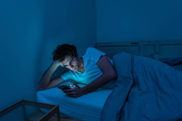 Addicted man chatting and surfing on the Internet with smart phone late at night in bed. Bored, sleepless and tired in dark room with moody light. In insomnia and mobile addiction concept. - Фото, изображение