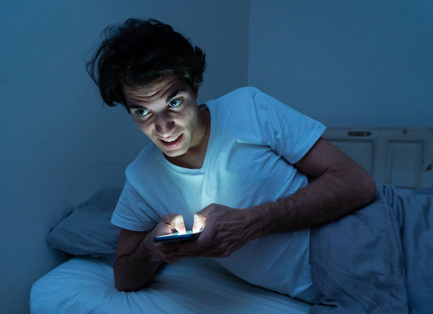 Addicted man chatting and surfing on the Internet with smart phone late at night in bed. Bored, sleepless and tired in dark room with moody light. In insomnia and mobile addiction concept. - Foto, imagen