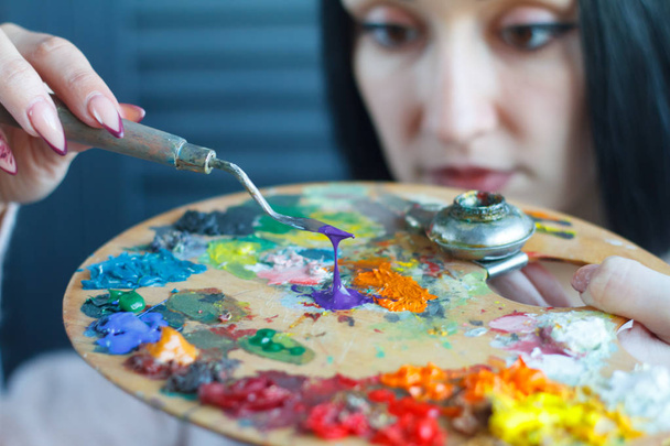 Closeup of a young woman with black hair mixes paint on a palette with a spatula against a white canvas, creating an oil painting - Photo, Image