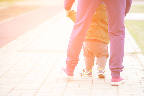 Lindo niño aprendiendo a caminar y dar sus primeros pasos. Mamá le sostiene la mano. niños pies de cerca, vista desde la espalda
 - Foto, imagen