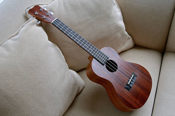 Um ukulele de madeira de quatro cordas é deixado apoiado verticalmente em um sofá branco
 - Foto, Imagem