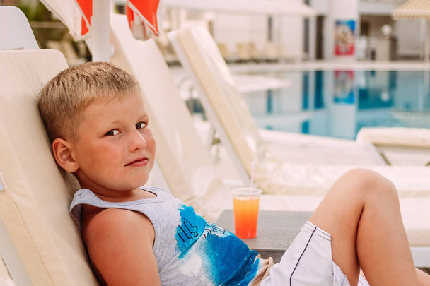 a cute blond Caucasian seven-year-old boy is lying by the outdoor pool. orange fruit cocktail . - Fotografie, Obrázek