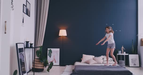 Litl girl Dancing on bed and listening to music with wireless headphones. Leisure time and modern lifestyle concept. Girl in pajama jumping on bed in morning and smiling. - Footage, Video