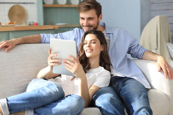 Young couple watching media content online in a tablet sitting on a sofa in the living room. - Zdjęcie, obraz