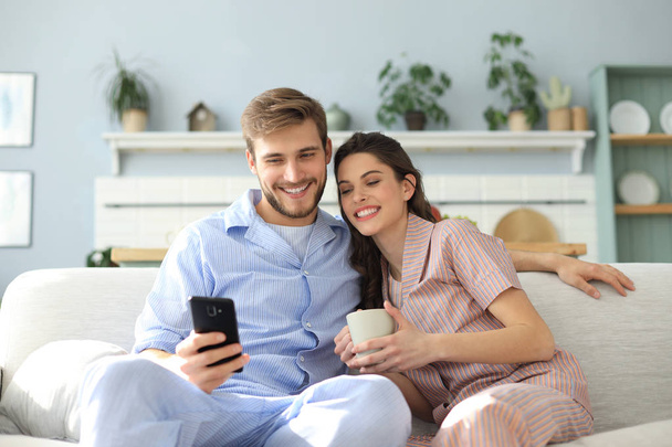 Giovane coppia che guarda contenuti online in uno smartphone seduto su un divano a casa in soggiorno. - Foto, immagini