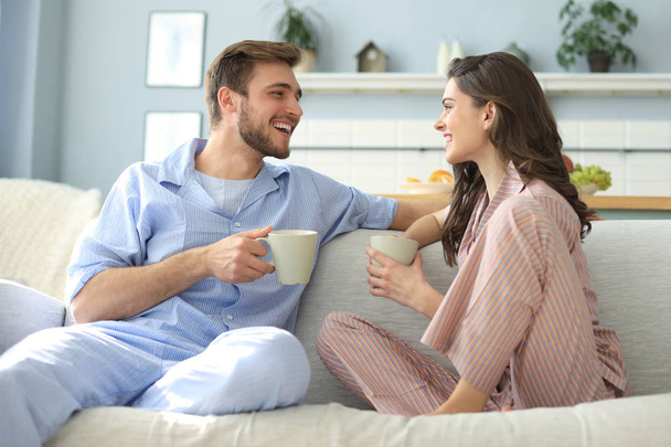 Beautiful young couple in pajamas is looking at each other and smiling on a sofa in the living room. - Foto, Bild