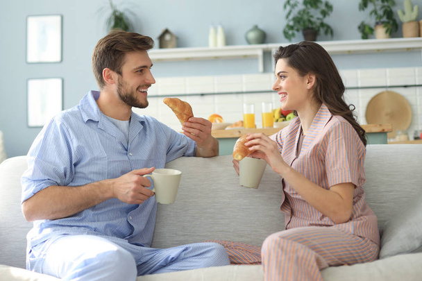 Happy young couple in pajamas in kitchen having breakfast, feeding each other a croissant. - Foto, Imagen