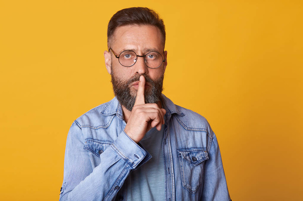Serious middle aged emotionless bearded man in blue shirt, showing silence gesture, asks to keep secret, putting forefinger on lips, male dressed in denim jacket stands over pink background. - Valokuva, kuva