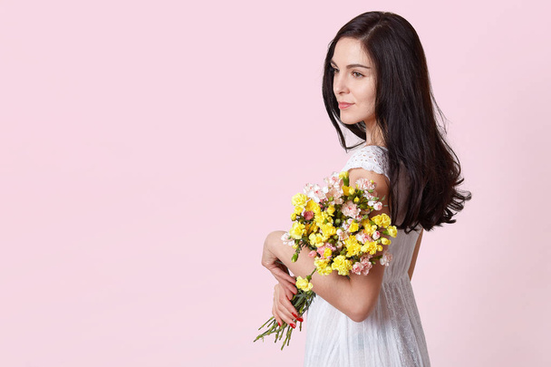 Half length portrait of charming brunette woman in white sundress holding bouquet of flowers over pink background, looking pensivly aside, copy space for your promotion content or advertisment. - Photo, image