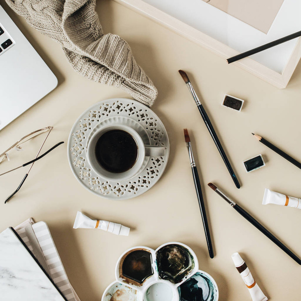 Watercolor painter home office desk workspace on beige background. Coffee cup, laptop, plaid, paint brushes, painting, watercolor. Flat lay, top view art blog, social media, website concept composition. - 写真・画像