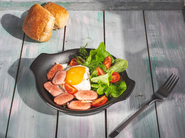 Petit déjeuner d'œufs brouillés avec saucisses, tomates et laitue dans une poêle noire. Servir sur un plateau à planches en bois avec petits pains
. - Photo, image