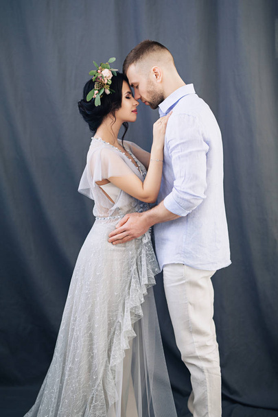 Pregnant european woman with her husband on gray background, young european couple waiting for a child, prenant woman with black hair in long light dress with wreath of flowers, happy european future - Foto, imagen