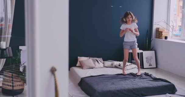 Litl girl Dancing on bed and listening to music with wireless headphones. Leisure time and modern lifestyle concept. Girl in pajama jumping on bed in morning and smiling. - Video