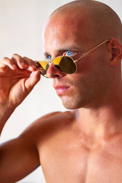 青い目とセクシーな筋肉質の美しい男性は、裸の胴体とデニムジーンズと金のサングラスを着用します。健康的な日焼けポーズを持つフィットネス男性. - 写真・画像