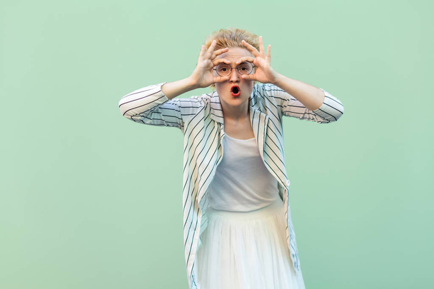 surprised young blonde woman in white striped blouse with eyeglasses standing with with binoculars hands gesture on eye in green background. - Photo, image
