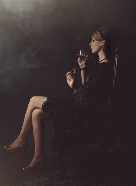 Een meisje in een zwarte jurk zit op een stoel met een glas wijn - Foto, afbeelding