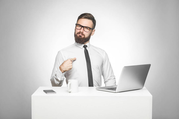 satisfied confident smiling bearded young manager in white shirt and black tie pointing himself and looking at camera while sitting at office table with laptop  - Фото, изображение
