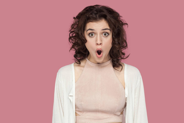surprised brunette young woman with curly hairstyle in casual style standing with shocked face and looking at camera with big eyes and open mouth on pink background. - 写真・画像