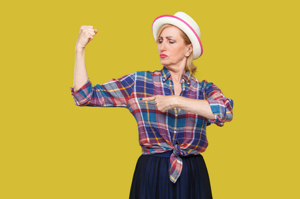 proud modern stylish mature woman in casual style with white hat standing and pointing finger at her biceps on yellow background. - Foto, Bild