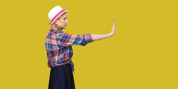 side view of serious modern stylish mature woman in casual style with hat looking forward and showing stop sign on yellow background. - Foto, Imagen