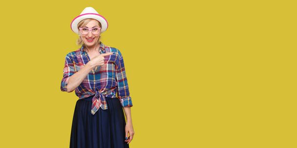 Portrait of happy modern stylish mature woman in casual style with hat and eyeglasses looking at camera and pointing aside on yellow background - Fotografie, Obrázek