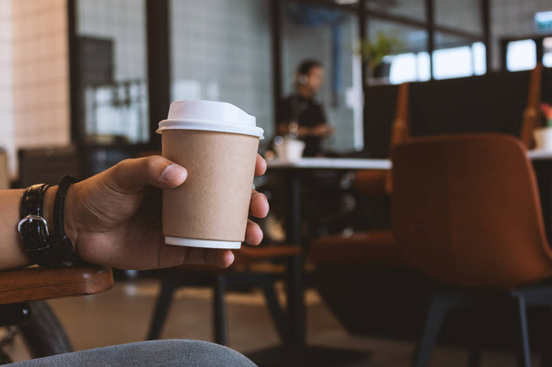 Рука крупным планом держит бумажную чашку кофе в кафе с мягким фокусом и над светом на заднем плане - Фото, изображение