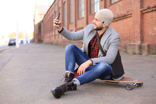 Handsome young man in grey coat and hat sitting on the longboard and taking selfie on the street in the city. - Foto, Imagem