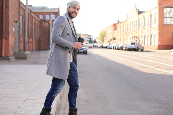 Όμορφος νέος άντρας με γκρι παλτό και καπέλο που διασχίζει το δρόμο με ένα φλιτζάνι καφέ. - Φωτογραφία, εικόνα