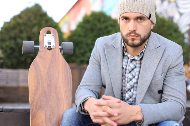 Handsome young man in grey coat and hat, resting, sitting with longboard. Urban skateboarding concept. - Zdjęcie, obraz