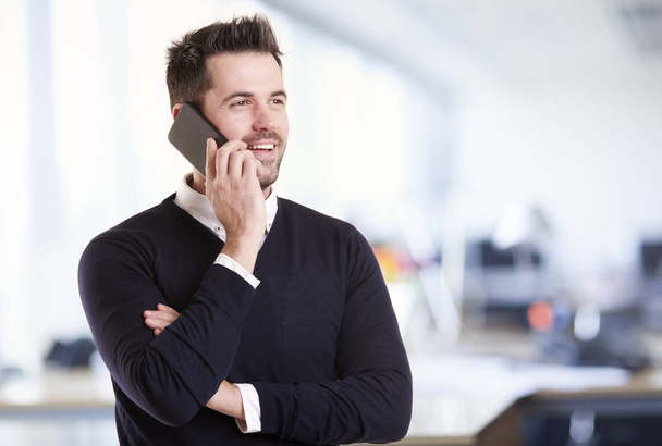 Πορτρέτο του όμορφου επιχειρηματία που στέκεται στο γραφείο και χρησιμοποιεί το κινητό του τηλέφωνο ενώ κάνει μια κλήση. - Φωτογραφία, εικόνα