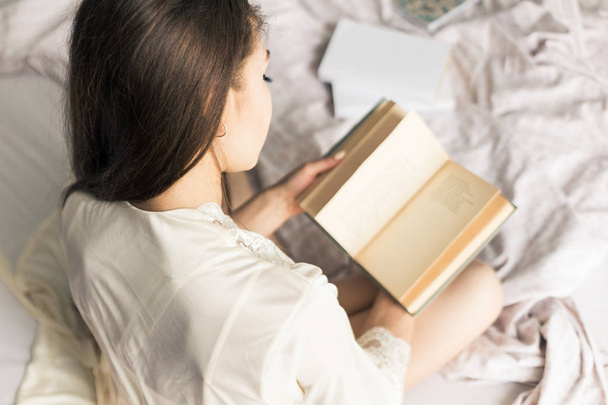 Atractiva joven en un camisón se sienta en la cama y lee un libro
 - Foto, Imagen