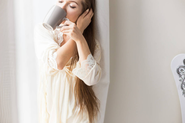Ελκυστική νεαρή κοπέλα με πυτζάμες στέκεται στο παράθυρο και πίνει καφέ. Πρωινή διάθεση - Φωτογραφία, εικόνα