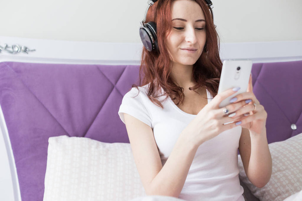 Attractive young brunette woman sits in the morning in her bed with headphones and a smartphone and listens to music in a good mood - Fotografie, Obrázek
