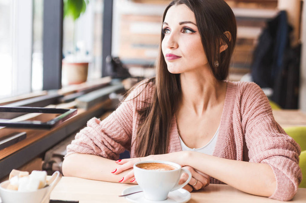 Όμορφη νεαρή γυναίκα κάθεται σε ένα τραπέζι σε ένα καφέ με ένα φλιτζάνι καφέ και κοιτάζει έξω από το παράθυρο - Φωτογραφία, εικόνα