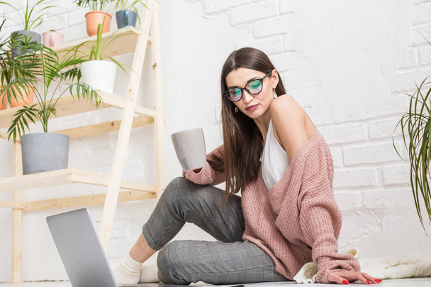 A young happy woman sits on the floor in a bright apartment or office interior and works at a laptop, freelancer girl at work, the concept of distance learning students, paperwork and online learning - Foto, Imagem