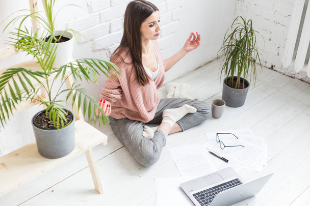 Young happy woman sits on the floor in a yoga pose in a bright apartment and works behind a laptop, a freelancer girl meditates while working. Stress of work - Фото, изображение
