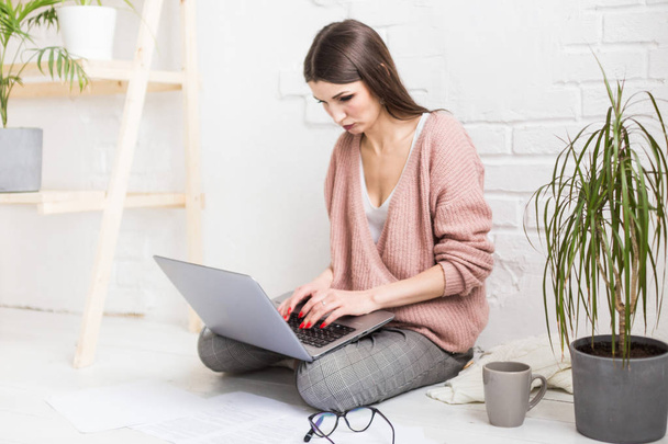 A young happy woman sits on the floor in a bright apartment or office interior and works at a laptop, freelancer girl at work, the concept of distance learning students, paperwork and online learning - 写真・画像