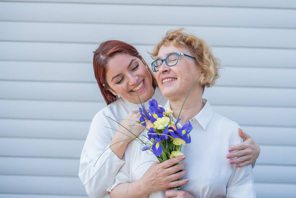 Adult daughter gives flowers to her mother outside, in the courtyard of the house. Spending time together, celebrating at home on weekends. Mothers Day. Warm intergenerational relationships - Фото, изображение