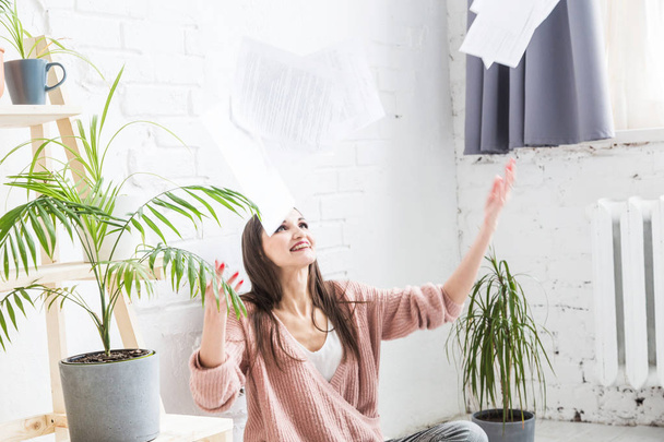 A young happy woman throws up a bunch of pieces of paper and documents sitting near the laptop, the freelance girl is happy and in a good mood. excitement and inspiration - Foto, Imagem