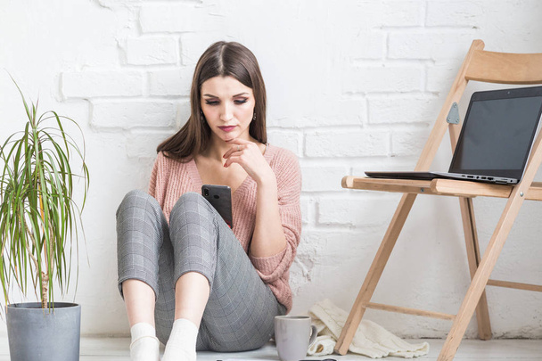 A happy woman in homemade casual clothes sits in a bright interior and uses a telephone, the girl is resting while working at a laptop and surfing the Internet. Cozy freelance work space, - Фото, зображення