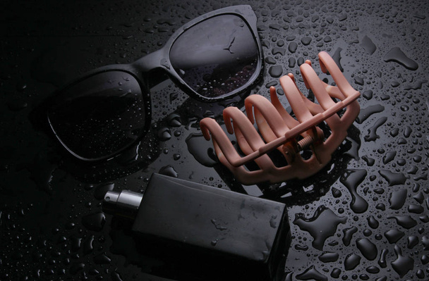 Gafas de sol, clip para el cabello, botella de perfume sobre fondo negro con gotas de agua. Moda cóncava
 - Foto, Imagen