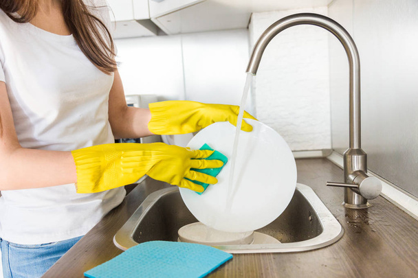 Eine junge Frau in gelben Handschuhen spült Geschirr mit einem Schwamm in der Spüle. Professioneller Reinigungsservice. - Foto, Bild