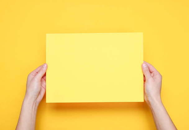 Mãos femininas segurando folha de papel amarelo no fundo amarelo. Vista superior. Mockup, espaço de cópia, minimalismo
 - Foto, Imagem
