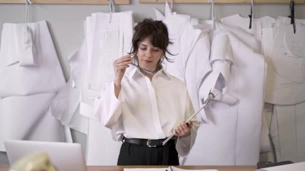 Middle shot of female fashion designer putting on eyeglasses and make notes - Кадри, відео