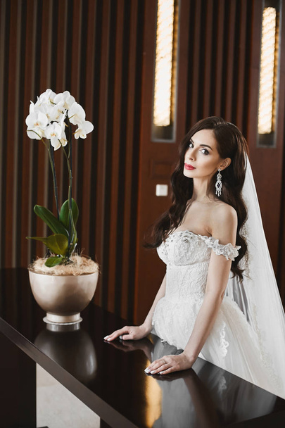 Young sexy bride with perfect body and big breasts in the fashionable wedding dress and in big luxury earrings posing at luxury interior - Foto, imagen