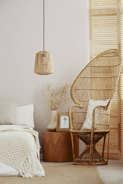 White and bright bedroom interior with wicker peacock chair with white pillow, rattan lamp and wooden nightstand with frame with poster and vase with flowers - Fotó, kép