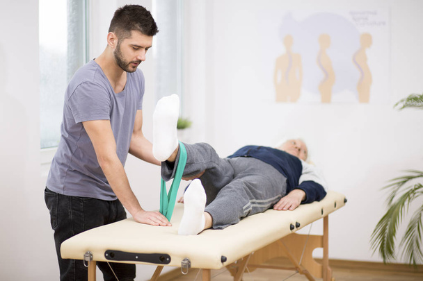 Grey elderly man lies on a physiotherapy table, and young doctor helps him during exercises - Foto, imagen
