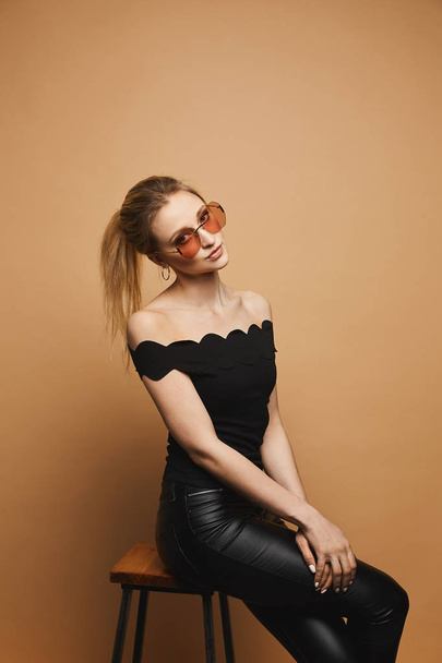 Young beautiful woman with slim perfect body in black leather pants, in a black blouse and in fashionable sunglasses at the orange background, isolated - Fotó, kép